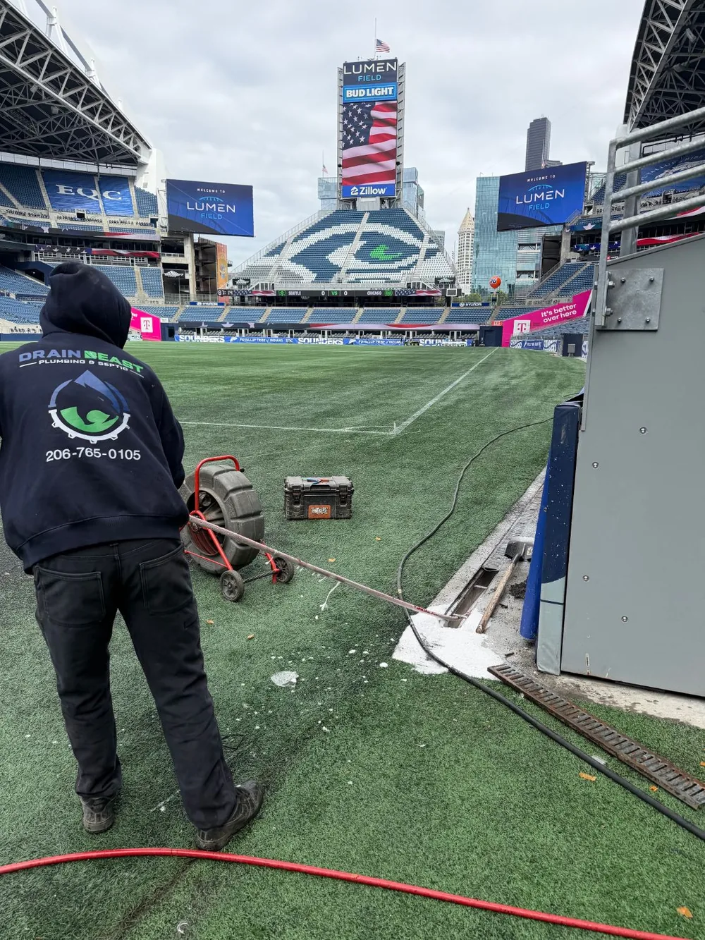 A technician in Drain Beast Services clothing on the touchline at Lumen Field, with the jetting reel set up on the turf.