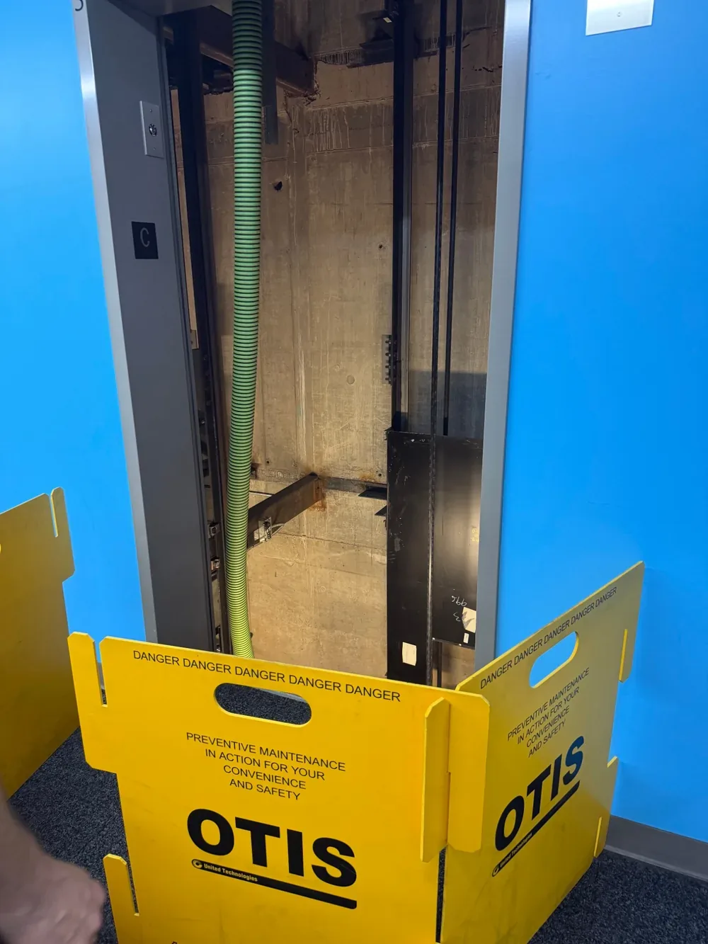 An elevator shaft with the doors open and a suction hose lowered into the pit, behind a maintenance barrier.