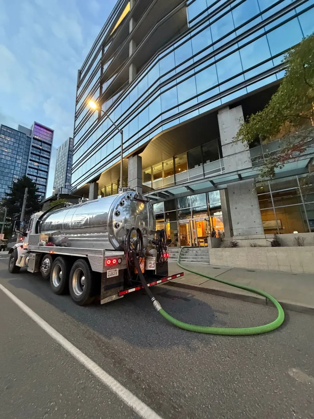 The vacuum truck parked on the street outside the building at dusk, with the suction hose running toward the entrance.