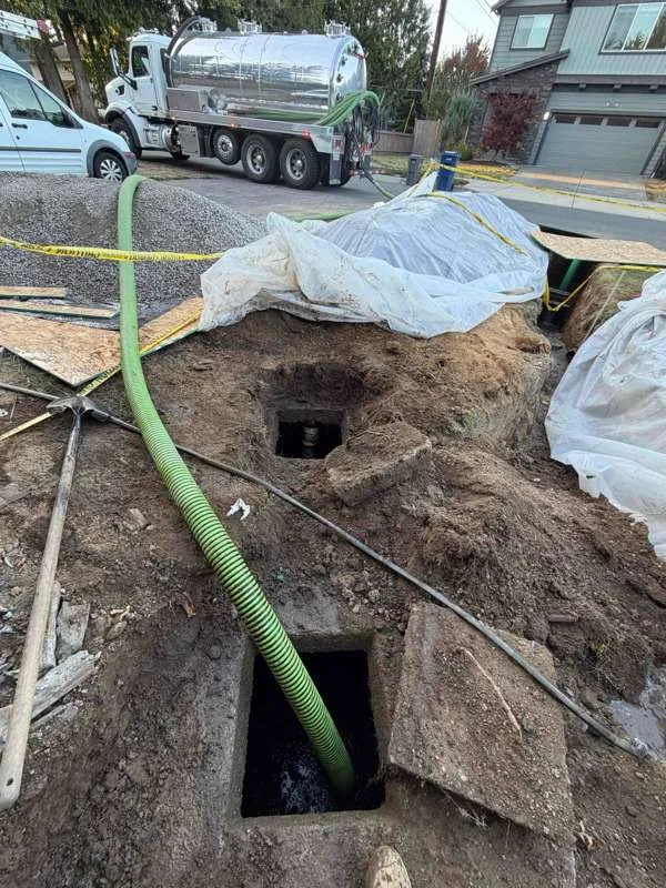 Both compartment openings uncovered in the excavation, with the suction hose run down to the vacuum truck on the street.