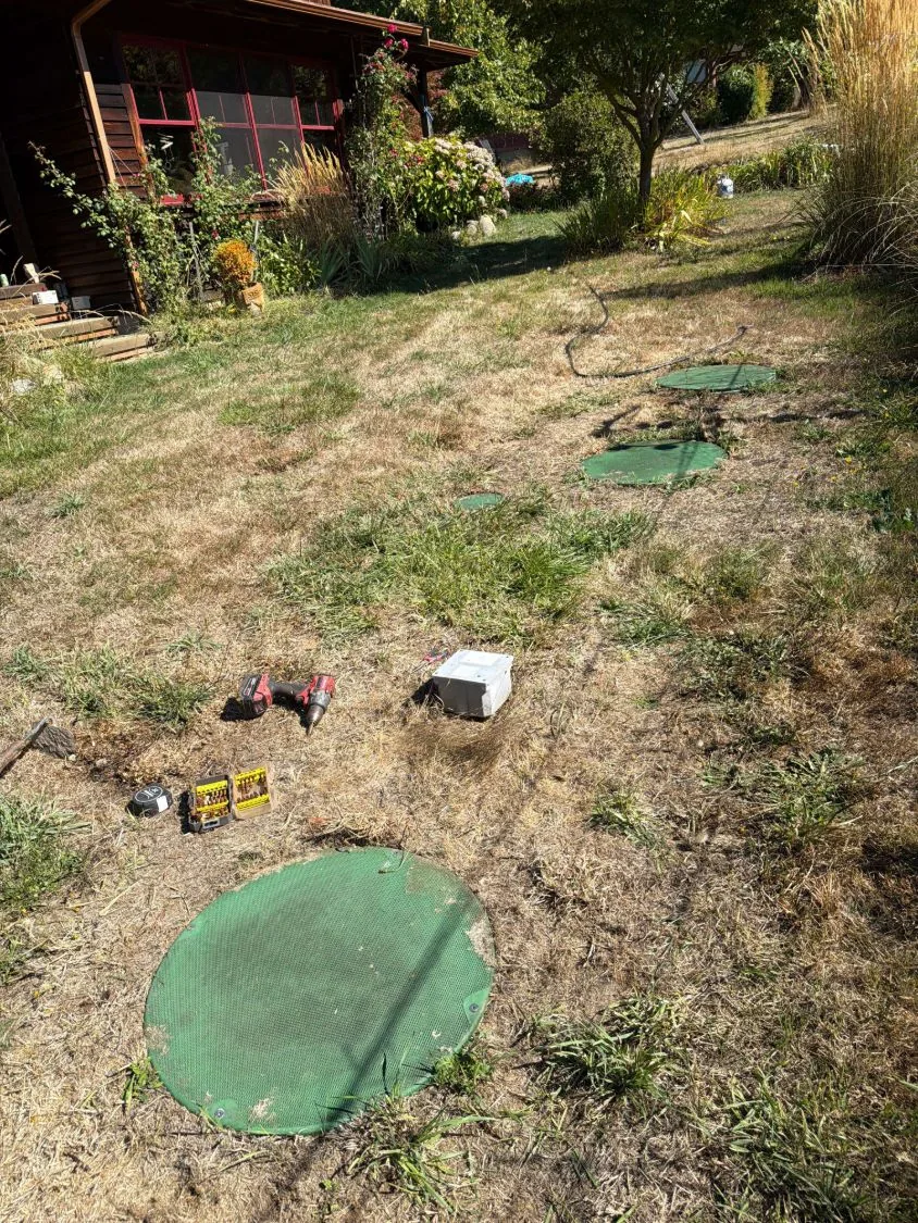 The system laid out across the garden: four riser lids, the technician's tools, and the junction box opened up on the grass.