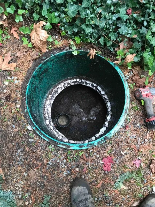The top of the tank exposed inside its riser, with a drill set on the ground beside the opening.
