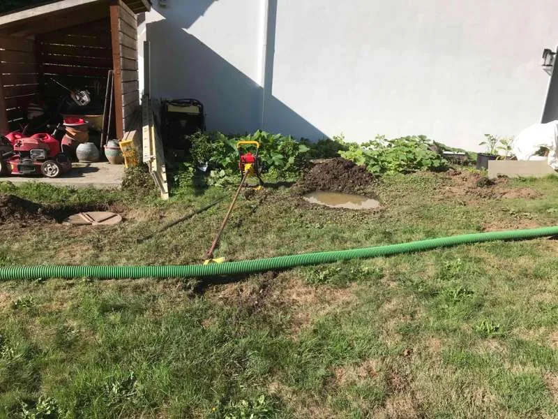 The work area in the back garden: the suction hose run across the lawn to the trash pump, with an exposed riser lid at the left and the excavated ground beside it.