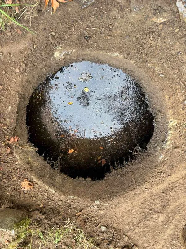 Looking down an excavated hole at a round tank opening filled to the brim with dark liquid.
