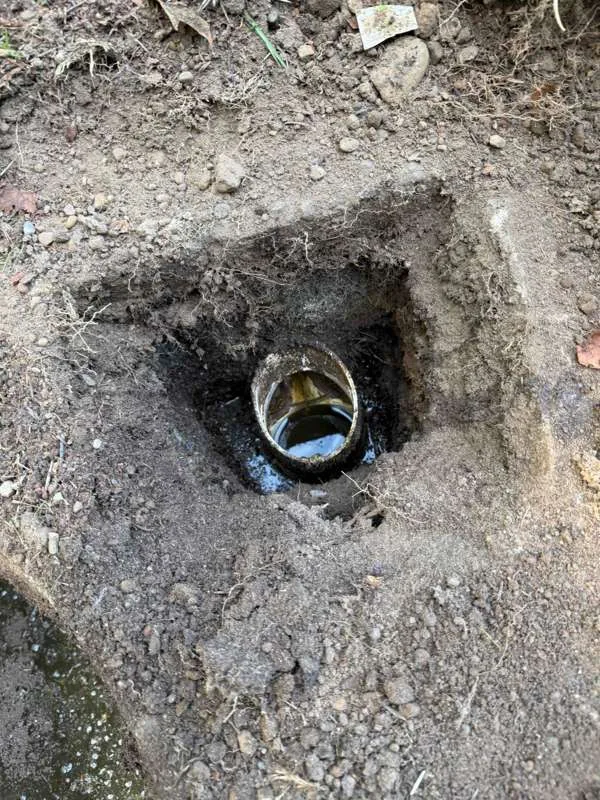 A pipe connection exposed in a hand-dug hole beside the tank, with liquid standing in the pipe.