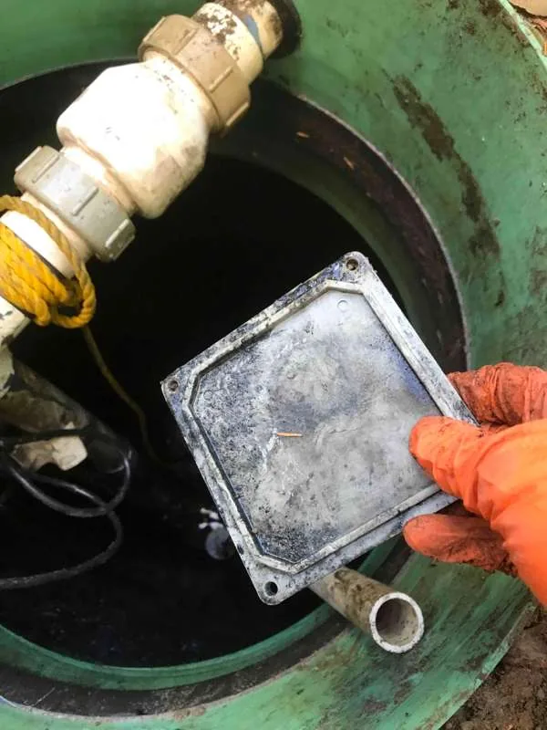 A gloved hand holding the junction box lid recovered from the bottom of the tank.
