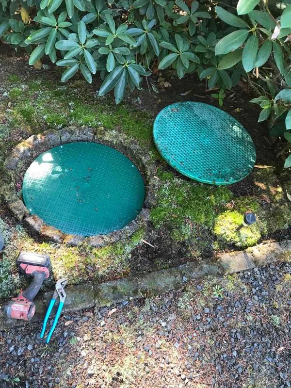Two green tank lids, one seated in its concrete riser and one lifted off and set on the ground beside it, with tools laid out on the gravel.